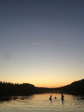 The silouettes of three People, who are Standing waist Deep in a lake. Before them lays a lies a dark forrest and a colourful sunset.